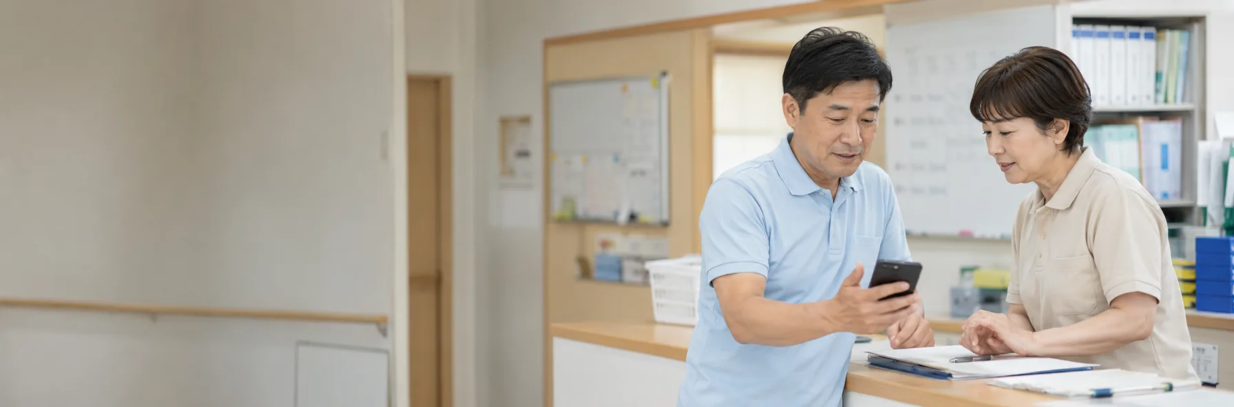 介護施設のスタッフステーションで、スマートフォンを見ながら引き継ぎをしている職員2名